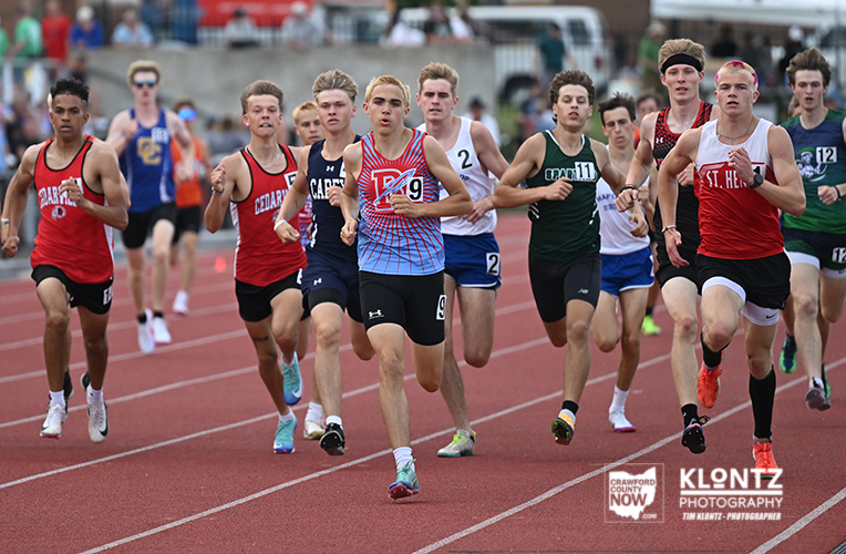 Group of male sprinters in a track race, wearing red, blue, green, and white uniforms, racing on a red track with bib numbers visible.