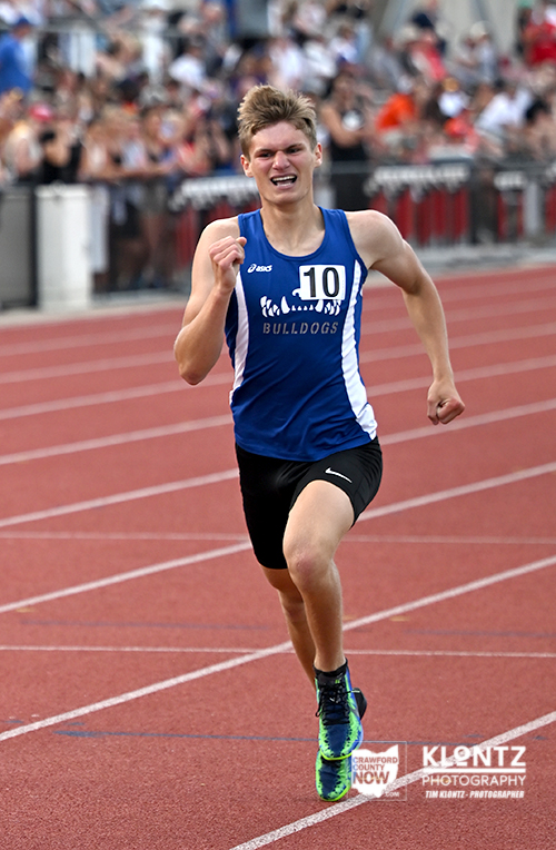 Male sprinter in a blue Bulldogs uniform with bib 10 sprinting on a red track, crowd cheering in the background.