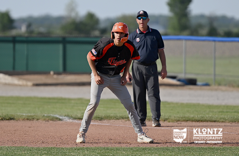 Baseball player in orange helmet and black jersey on infield, ready to run, coached by a man in the background.