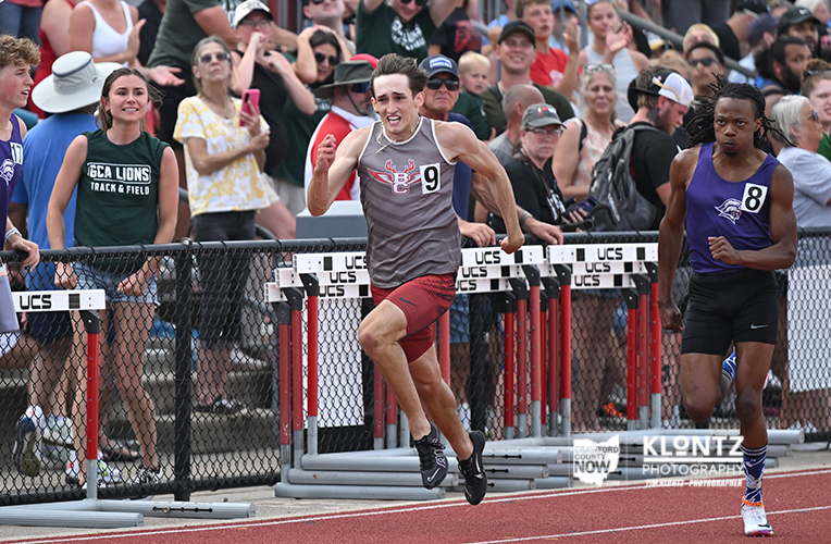 Male track athlete in gray tank top and red shorts sprinting over hurdles during a race, number 9, with a purple-jersey runner nearby and a crowded stadium in the background.