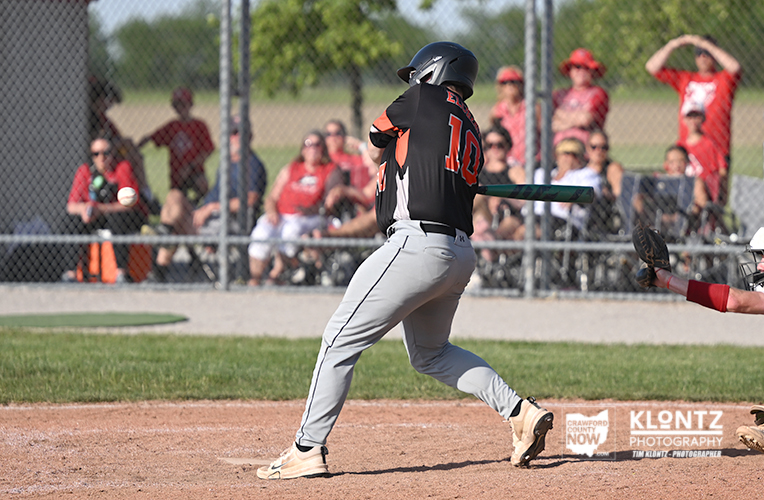 Baseball batter in black jersey swinging at a pitch, number 12, at home plate with catcher in red gear nearby and a chain-link fence in the background.