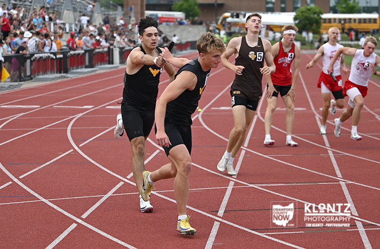 Group of male sprinters racing on a red track, wearing black, red, and white uniforms, with a crowd along the sidelines.