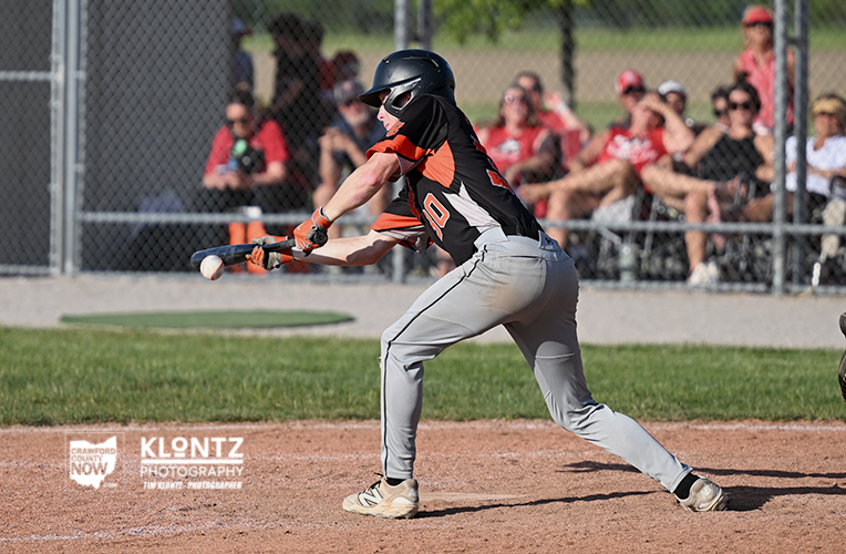 A baseball batter in black and orange swings at a pitch on a dirt field, crowd watching behind a chain-link fence.