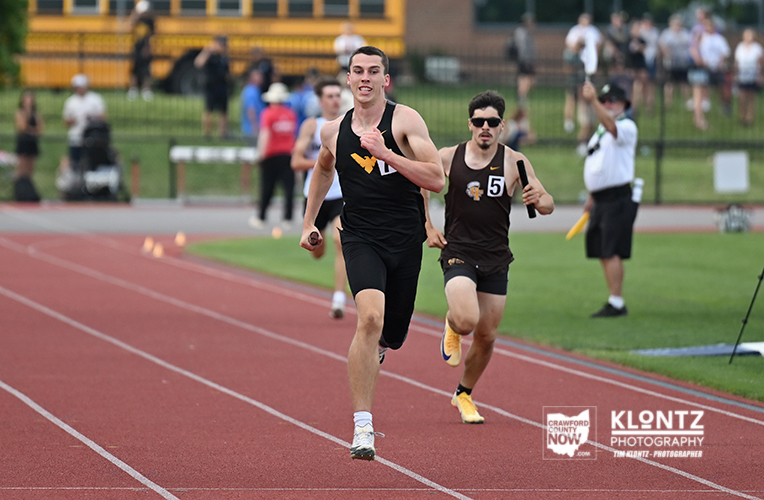 Two male sprinters race on a red track at a track meet; lead runner wears a black singlet with white shoes, followed by a competitor in brown with yellow shoes, with spectators in the background.
