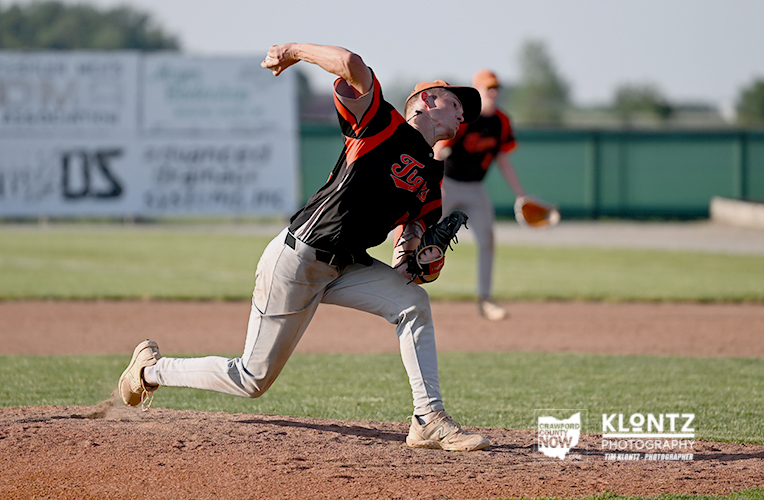 Baseball pitcher in a black-orange uniform mid-throw on the mound, with a glove up and leg forward as a teammate watches in the background.
