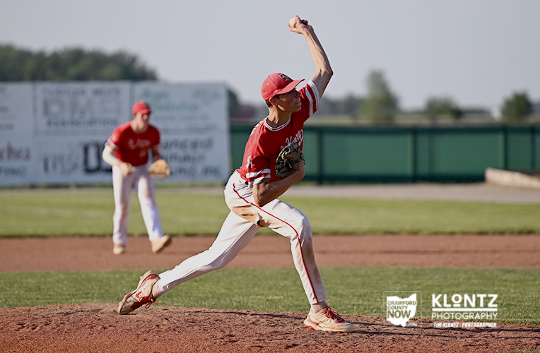 Baseball pitcher in red uniform throwing a pitch from the mound with a teammate in red infield behind him.