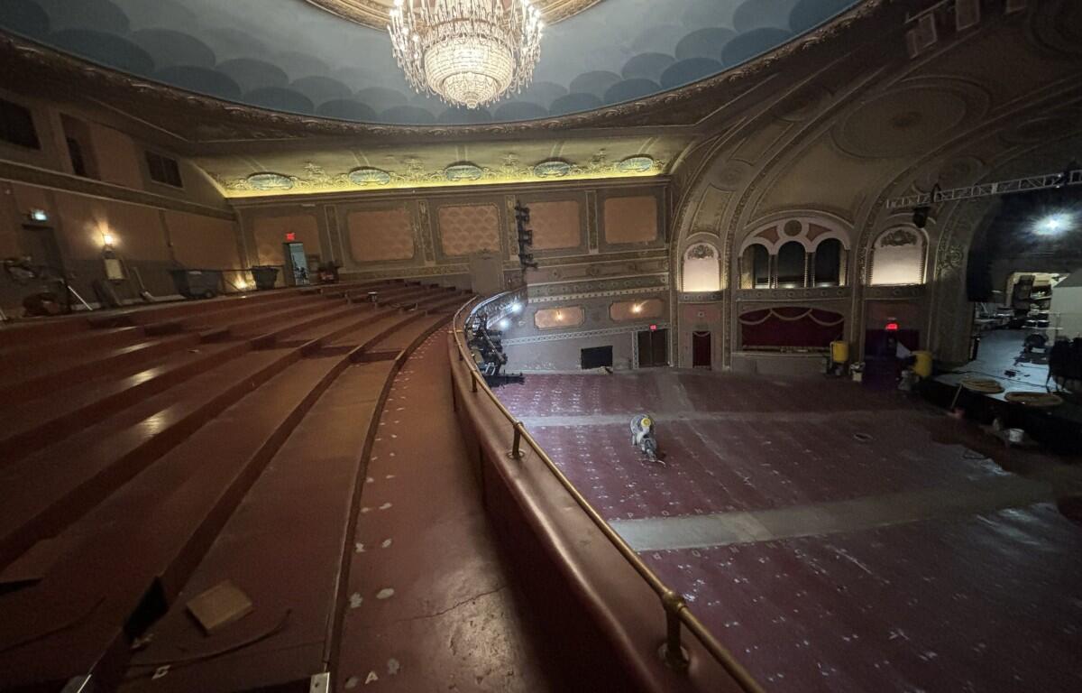 Ornate historic theater auditorium with a large chandelier, empty red seats, and renovation equipment on the stage area.