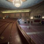 Ornate historic theater auditorium with a large chandelier, empty red seats, and renovation equipment on the stage area.