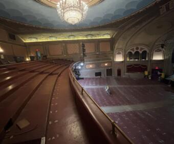 Ornate historic theater auditorium with a large chandelier, empty red seats, and renovation equipment on the stage area.