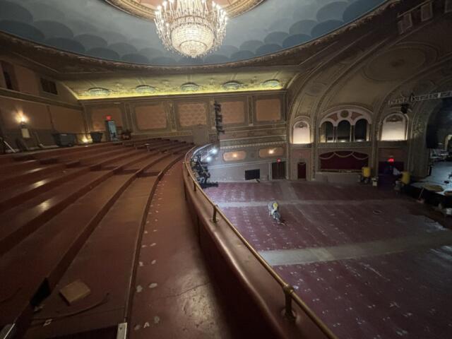 Ornate historic theater auditorium with a large chandelier, empty red seats, and renovation equipment on the stage area.