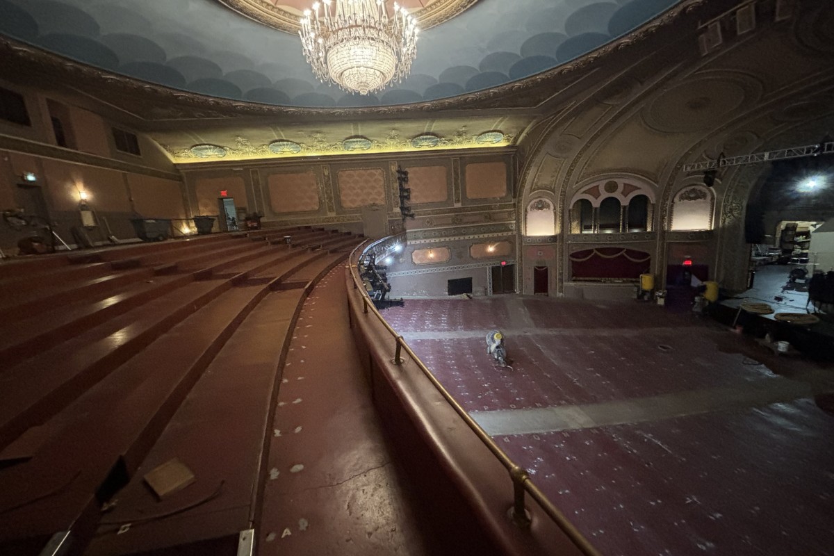 Ornate historic theater auditorium with a large chandelier, empty red seats, and renovation equipment on the stage area.