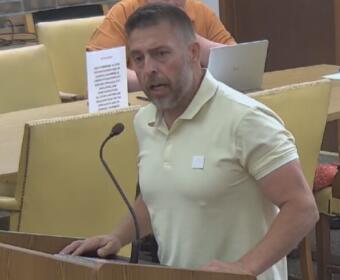 Man in a pale yellow polo shirt speaks at a wooden podium with a microphone in a meeting room, a laptop and water bottle visible behind him