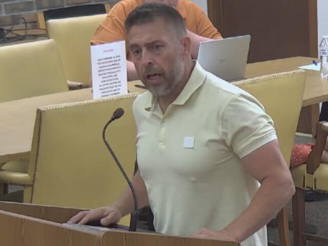 Man in a pale yellow polo shirt speaks at a wooden podium with a microphone in a meeting room, a laptop and water bottle visible behind him