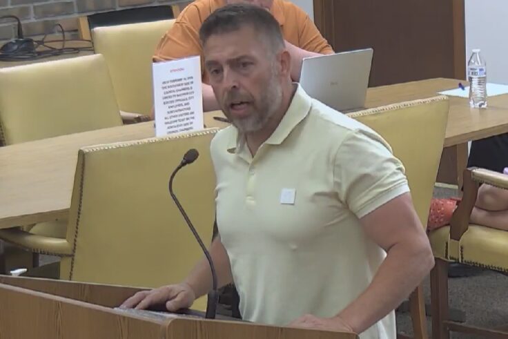 Man in a pale yellow polo shirt speaks at a wooden podium with a microphone in a meeting room, a laptop and water bottle visible behind him