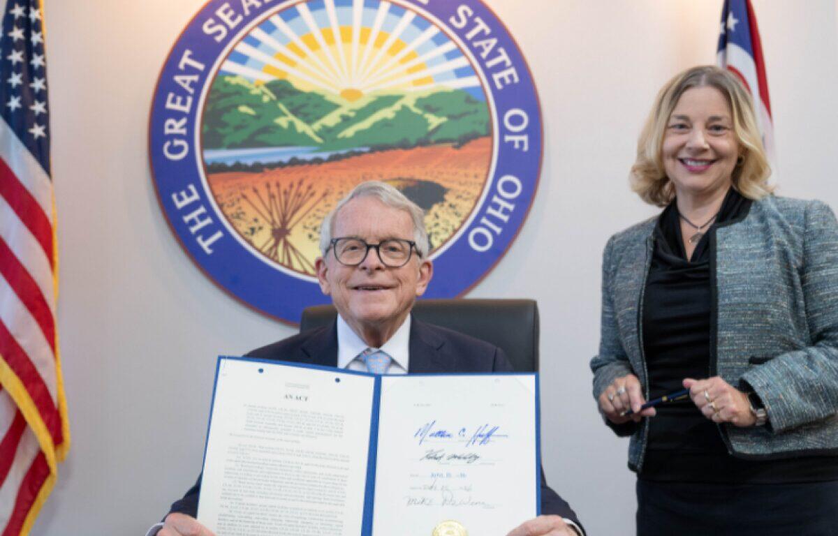 Man in a suit seated, holding an open signed document while a smiling woman stands beside him, with the Ohio Great Seal and flags in the background.