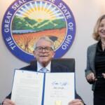 Man in a suit seated, holding an open signed document while a smiling woman stands beside him, with the Ohio Great Seal and flags in the background.