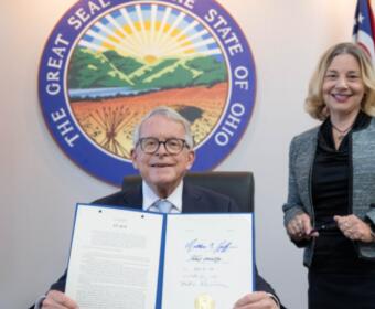 Man in a suit seated, holding an open signed document while a smiling woman stands beside him, with the Ohio Great Seal and flags in the background.