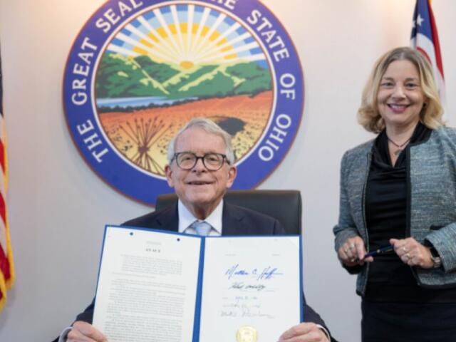 Man in a suit seated, holding an open signed document while a smiling woman stands beside him, with the Ohio Great Seal and flags in the background.