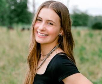 Smiling young woman with long light brown hair wearing a black top, standing in a grassy outdoor field with greenery behind her.