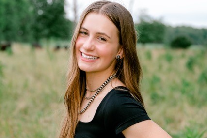 Smiling young woman with long light brown hair wearing a black top, standing in a grassy outdoor field with greenery behind her.