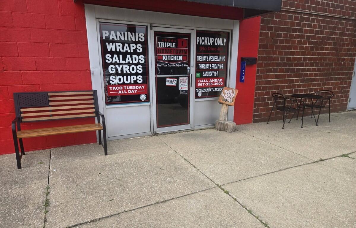 storefront with red wall and glass doors advertising paninis, wraps, salads, gyros and soups; bench on left and outdoor table with chairs on the right on a concrete sidewalk