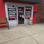storefront with red wall and glass doors advertising paninis, wraps, salads, gyros and soups; bench on left and outdoor table with chairs on the right on a concrete sidewalk