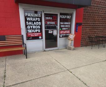 storefront with red wall and glass doors advertising paninis, wraps, salads, gyros and soups; bench on left and outdoor table with chairs on the right on a concrete sidewalk