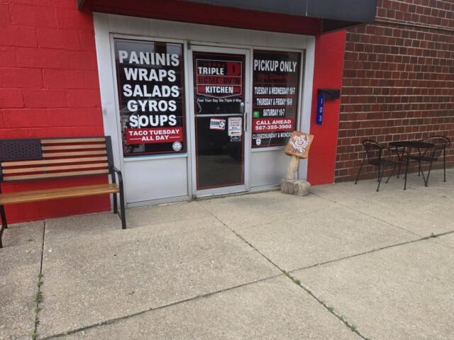 storefront with red wall and glass doors advertising paninis, wraps, salads, gyros and soups; bench on left and outdoor table with chairs on the right on a concrete sidewalk
