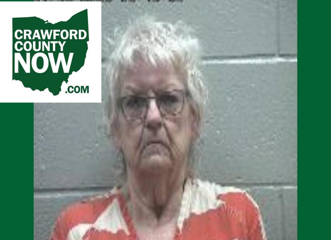 Mugshot-style photo of an elderly woman with short gray hair and glasses, wearing an orange and cream patterned blouse, against a gray cinder-block wall; Crawford County Now logo in the top-left corner.