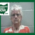 Mugshot-style photo of an elderly woman with short gray hair and glasses, wearing an orange and cream patterned blouse, against a gray cinder-block wall; Crawford County Now logo in the top-left corner.