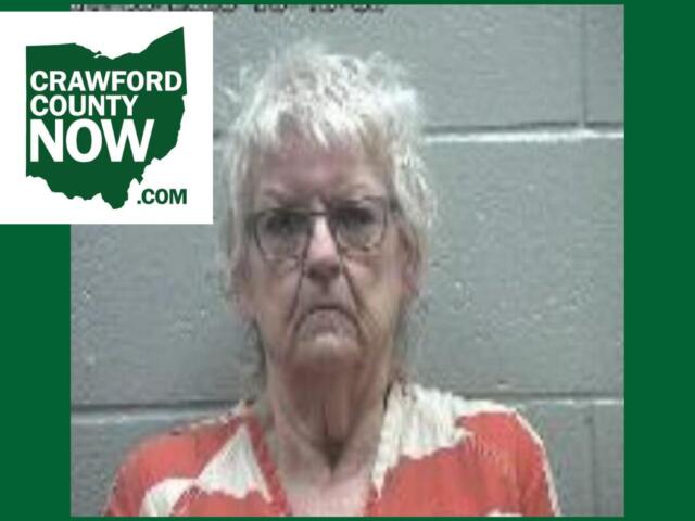 Mugshot-style photo of an elderly woman with short gray hair and glasses, wearing an orange and cream patterned blouse, against a gray cinder-block wall; Crawford County Now logo in the top-left corner.
