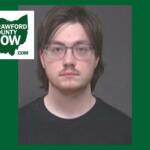 Mugshot-style portrait of a young man with glasses, dark hair, and a black shirt; Crawford County Now logo overlaps the image.