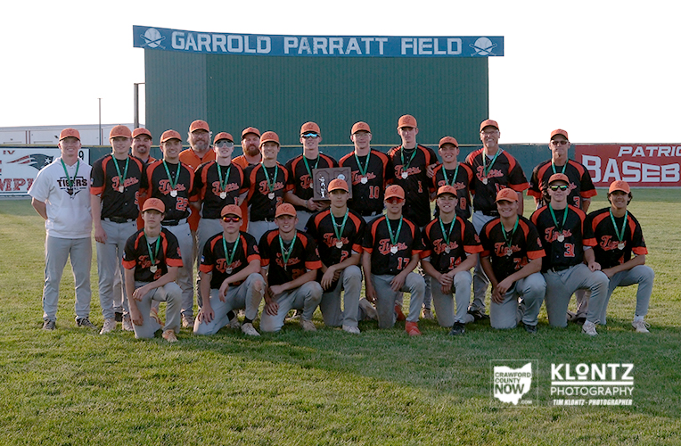 Baseball team in black and orange uniforms posing on the field with medals after a game, under a sign that reads Garrold Parratt Field.