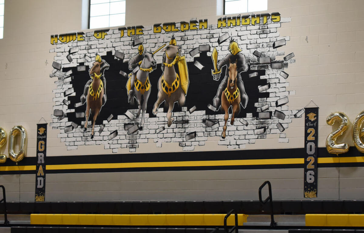 Gym mural 'Home of the Golden Knights' showing four armored knights breaking through a brick wall, with 2026 banners and gold 20/26 balloons.