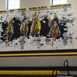 Gym mural 'Home of the Golden Knights' showing four armored knights breaking through a brick wall, with 2026 banners and gold 20/26 balloons.
