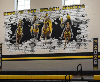 Gym mural 'Home of the Golden Knights' showing four armored knights breaking through a brick wall, with 2026 banners and gold 20/26 balloons.