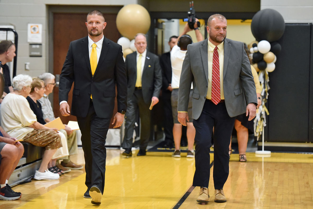 Two men in suits walk down a gymnasium aisle at a formal event, with seated spectators along the sides.