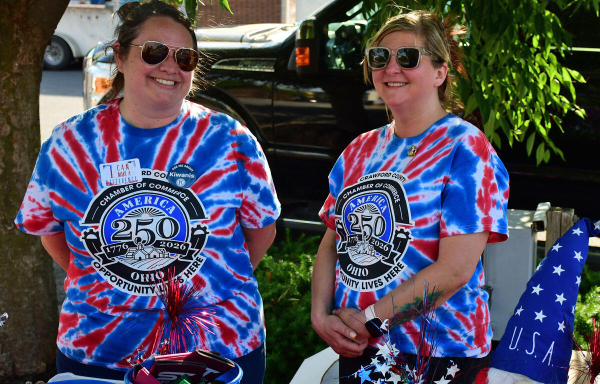 Two women in blue and red tie-dye shirts with a 250th anniversary emblem smile beside a car, with patriotic decorations nearby.