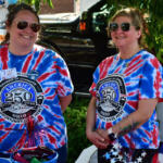 Two women in blue and red tie-dye shirts with a 250th anniversary emblem smile beside a car, with patriotic decorations nearby.
