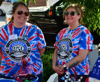 Two women in blue and red tie-dye shirts with a 250th anniversary emblem smile beside a car, with patriotic decorations nearby.