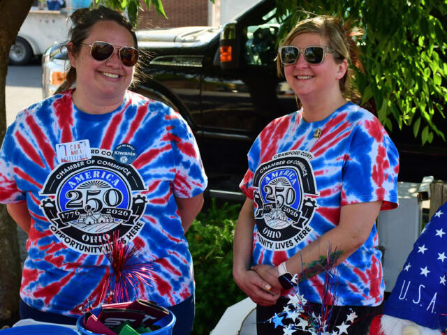 Two women in blue and red tie-dye shirts with a 250th anniversary emblem smile beside a car, with patriotic decorations nearby.