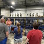 Instructor in an orange shirt explains machinery to a small group inside a factory, with blue industrial equipment behind a yellow safety barrier.