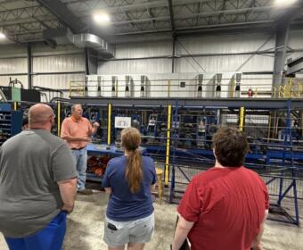 Instructor in an orange shirt explains machinery to a small group inside a factory, with blue industrial equipment behind a yellow safety barrier.