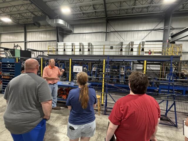 Instructor in an orange shirt explains machinery to a small group inside a factory, with blue industrial equipment behind a yellow safety barrier.