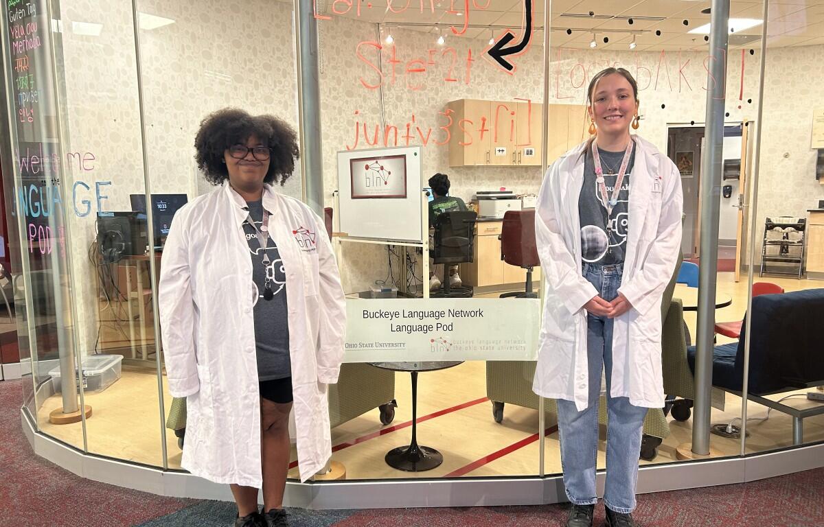Two students in white lab coats stand at a glass-walled language pod in a university lobby, smiling at the camera; sign reads Buckeye Language Network Language Pod.