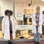 Two students in white lab coats stand at a glass-walled language pod in a university lobby, smiling at the camera; sign reads Buckeye Language Network Language Pod.