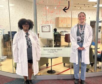 Two students in white lab coats stand at a glass-walled language pod in a university lobby, smiling at the camera; sign reads Buckeye Language Network Language Pod.
