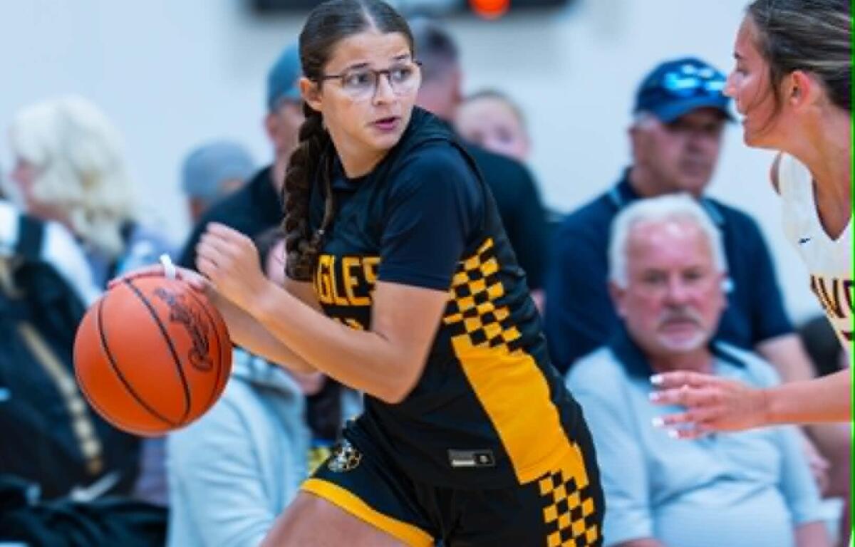 Female basketball player in a black and yellow checkered uniform dribbles an orange basketball, with a defender nearby and spectators in the background.