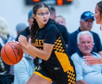 Female basketball player in a black and yellow checkered uniform dribbles an orange basketball, with a defender nearby and spectators in the background.
