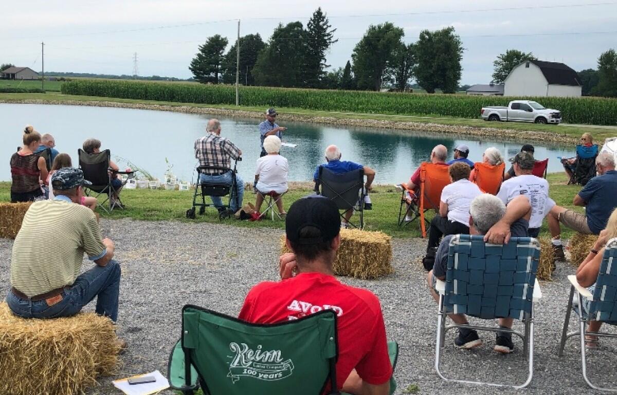 Group of people seated on hay bales and lawn chairs along a pond, listening to a man speaking at an outdoor gathering on a rural, sunny day.